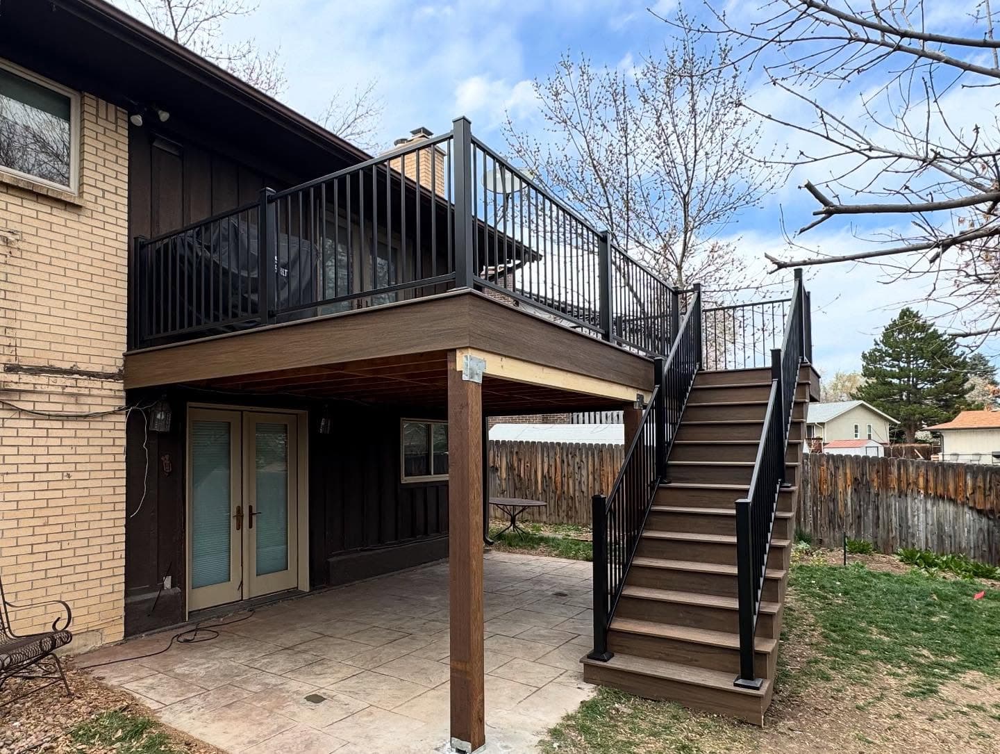 Elevated deck off a brick home with dark composite decking, black railing, and stairs to a stamped concrete patio